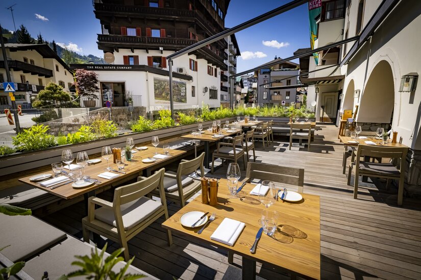 Außenterrasse eines Restaurants mit eingedeckten Holztischen, Stühlen und Blick auf alpine Gebäude.