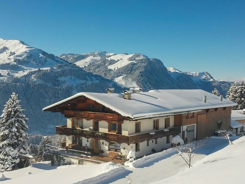 Hof Hamoos im Winter Ein schneebedeckter Bauernhof mit Holzbalkonen vor einer alpinen Bergkulisse im Winter.