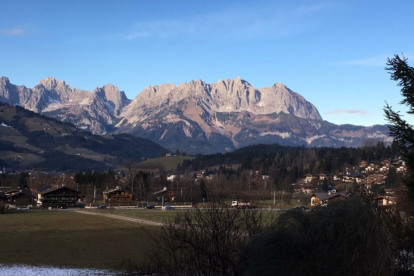 Ausblick auf den Wilden Kaiser Blick über ein Tal mit Häusern und Wiesen auf ein markantes, felsiges Gebirgsmassiv unter blauem Himmel.