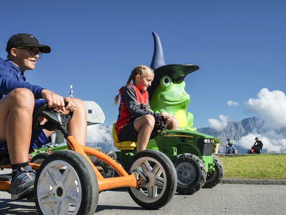 Zwei Kinder fahren mit kleinen Fahrzeugen auf einer sonnigen Wiese. Im Hintergrund ist eine große grüne Figur mit einem Zauberhut zu sehen.