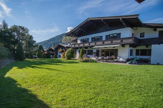 Landhaus Gasteiger im Sommer Alpines Landhaus mit großer Rasenfläche, Holzbalkonen und Terrasse unter blauem Himmel.