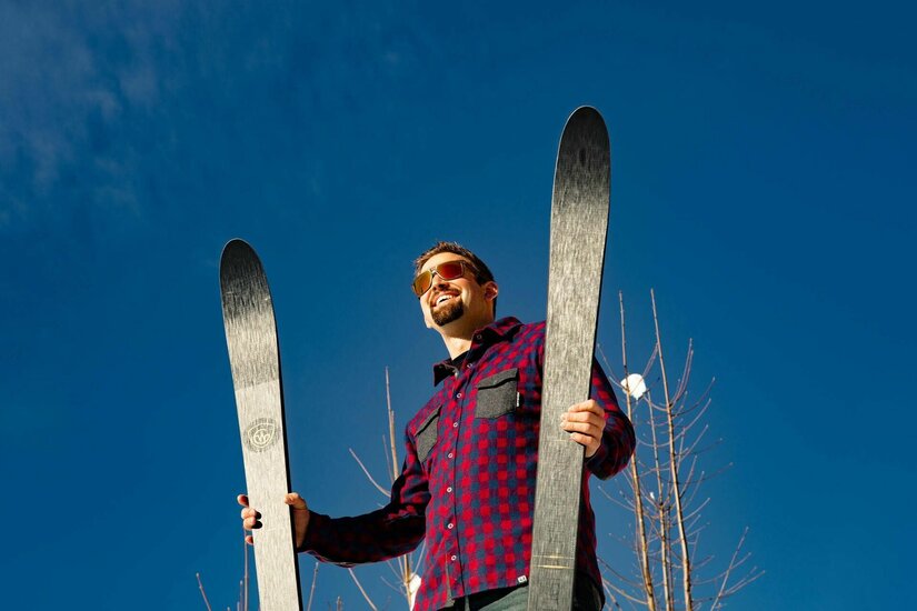 Skifahrer mit Ausrüstung Ein Mann im karierten Hemd hält zwei dunkle Skier senkrecht vor blauem Himmel.