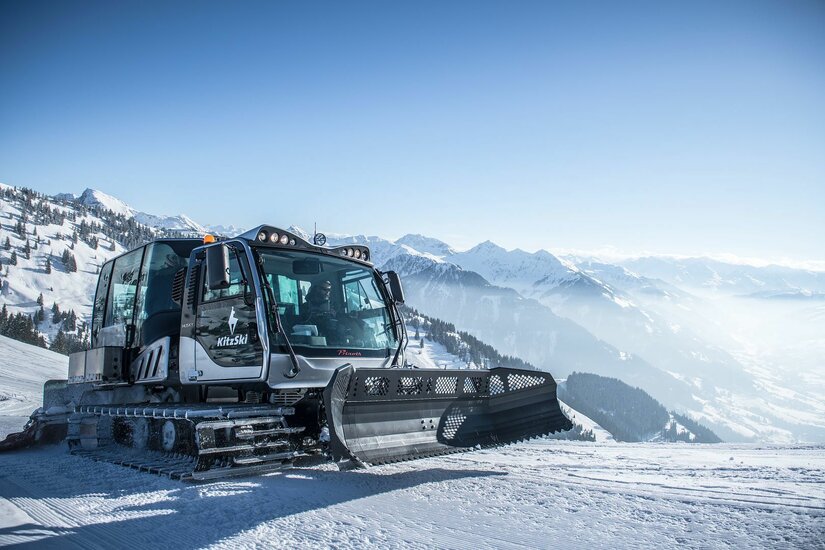 Pistenraupe im Skigebiet Eine silberne Pistenraupe mit KitzSki-Logo steht auf einer verschneiten Skipiste vor einem Bergpanorama.