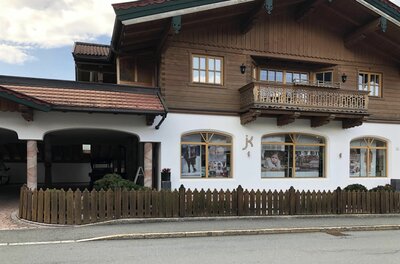 Zweistöckiges Gebäude im alpinen Stil mit Holzobergeschoss, Balkon und Schaufenstern hinter einem Holzzaun.