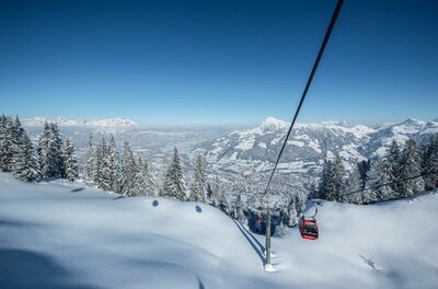Winterliches Skigebiet Kitzbühel Rote Gondel einer Seilbahn über verschneiten Hängen und Tannen vor Gebirgspanorama.