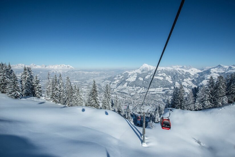 Winterliches Skigebiet Kitzbühel Rote Gondel einer Seilbahn über verschneiten Hängen und Tannen vor Gebirgspanorama.
