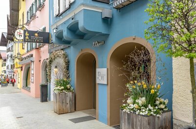 Blaue Hausfassade mit Rundbogen-Eingängen und hölzernen Blumenkästen in einer gepflasterten Fußgängerzone.