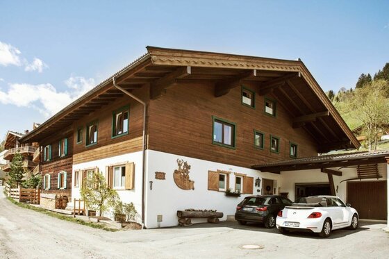 Zweistöckiges Alpenhaus mit weißer Fassade im Erdgeschoss, dunkler Holzverkleidung oben und geparkten Autos davor.
