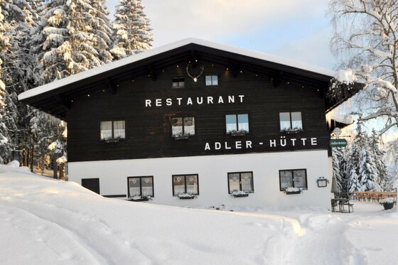 Vorderansicht der Adlerhütte im Schnee mit dunkler Holzfassade und weißem Erdgeschoss vor verschneiten Tannen.