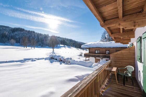 Holzbalkon mit Blick auf eine verschneite Wiese, Nadelwald und ein benachbartes Bauernhaus bei Sonnenschein.
