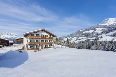 Ein traditionelles Alpenhaus mit Holzbalkonen in einer verschneiten Berglandschaft unter blauem Himmel.