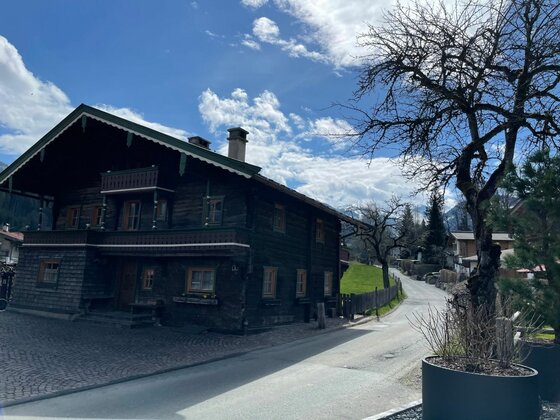 Holzhaus am Auracher Höhenweg Ein traditionelles dunkles Holzhaus mit Balkon an einer asphaltierten Straße unter blauem Himmel.