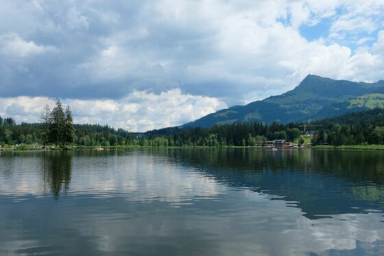 See mit Bergpanorama Blick über einen ruhigen See auf bewaldete Ufer und einen markanten Berggipfel unter bewölktem Himmel.