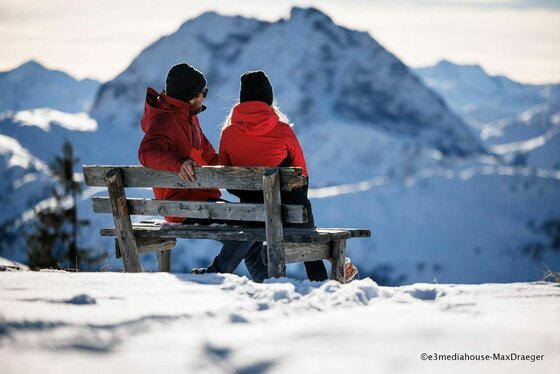 Winterrast auf der Holzbank Zwei Personen in roten Jacken sitzen auf einer Holzbank im Schnee vor einem Bergpanorama.
