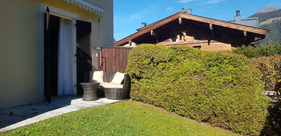 Sonnige Terrasse und Garten Terrasse mit Rattanmöbeln, kleiner Rasenfläche und Hecke vor einem Holzhaus.