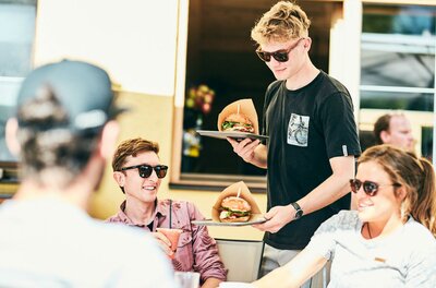 Simple food & drinks Ein Kellner bringt Burger zu einer Gruppe von Freunden in einem Restaurant. Alle tragen Sonnenbrillen und genießen die Atmosphäre.