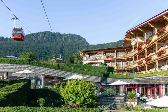 Blick auf das Hotel Kaiserhof in Kitzbühel mit der vorbeifahrenden roten Hahnenkammbahn vor bewaldeten Bergen.