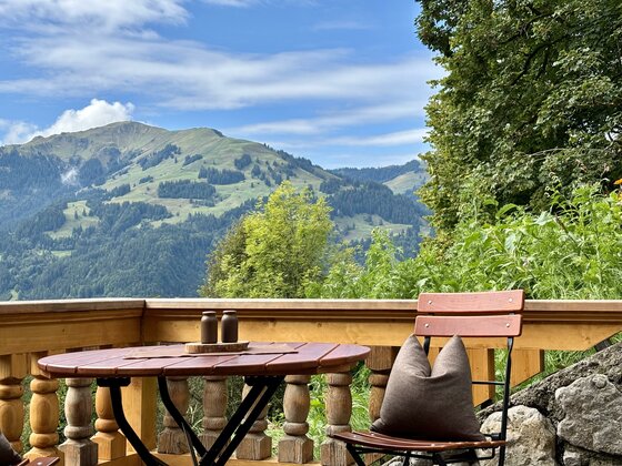 IMG_1892 2 Ein schöner Ausblick auf grüne Berge unter einem klaren Himmel. Auf der Terrasse stehen ein runder Tisch und ein Stuhl mit einem Kissen.
