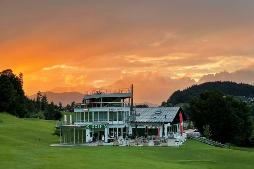 Modernes Zielhaus mit Restaurant auf einer grünen Wiese vor Gebirgskulisse bei Sonnenuntergang.