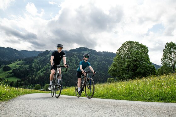 Rennradfahren im Frühling Zwei Personen auf Rennrädern fahren auf einer asphaltierten Straße neben einer Blumenwiese vor bewaldeten Bergen.