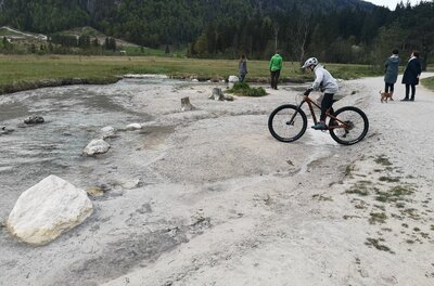 Ein Kind auf einem Mountainbike fährt an einem flachen Bachlauf vorbei, im Hintergrund Spaziergänger und Wald.