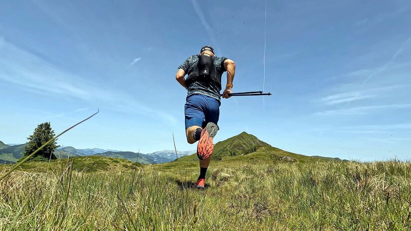 Rückansicht eines Trailrunners mit Laufweste und Stöcken auf einer Bergwiese.