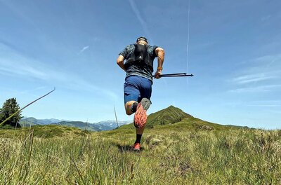 Trailrunning in den Bergen Rückansicht eines Trailrunners mit Laufweste und Stöcken auf einer Bergwiese.