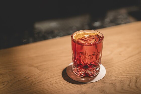 Getränk in der Mocking Kneipe Ein Glas mit rotem Getränk, Eiswürfeln und einer getrockneten Orangenscheibe auf einem Holztisch.