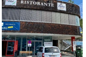 Behindertenparkplatz - Sportfeld 2a Ein Restaurant mit der Aufschrift "Ristorante" und einer Terrasse. Vor dem Gebäude steht ein weißes Auto auf einem Parkplatz.