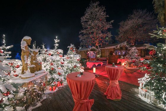 Weihnachtsmarkt bei Nacht mit beleuchteten Tannen, Stehtischen und Holzfiguren im Schnee.