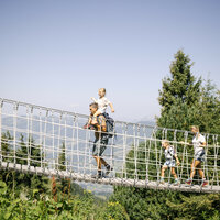 Eine Familie überquert eine schaukelnde Hängebrücke in den Bergen, während die Sonne am blauen Himmel scheint.