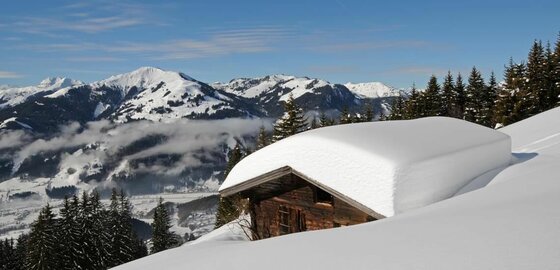 Winterliche Almhütte bei Jochberg Holzhütte mit dicker Schneeschicht auf dem Dach vor verschneitem Bergpanorama und Nadelwald.