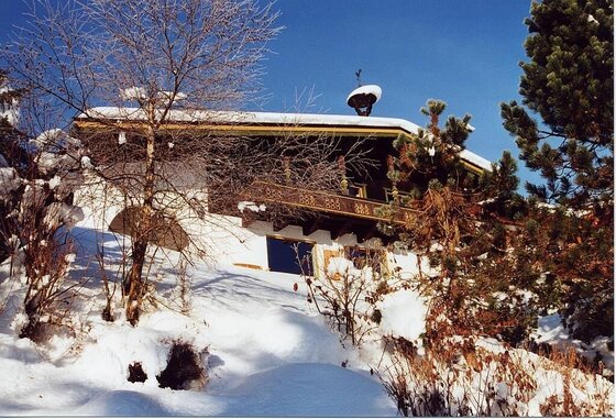 Landhaus Perktold im Winter Alpines Landhaus mit Holzbalustrade im verschneiten Hang unter blauem Himmel.