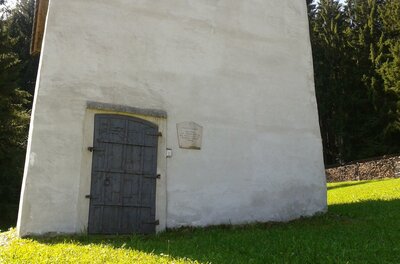 Ein schlichtes Gebäude mit einer dunklen Holztür steht auf grünem Gras. Im Hintergrund sind Bäume zu sehen.