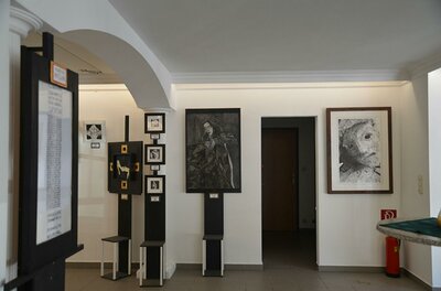 Ein moderner Ausstellungsraum mit mehreren Kunstwerken an den Wänden. Es gibt verschiedene Ständer für Bilder und Informationen.
