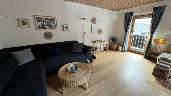 IMG_6491 Ein gemütliches Wohnzimmer mit einem dunklen Sofa und einem runden Tisch. Durch die Fenster scheint Tageslicht und es gibt einen Zugang zu einem Balkon.