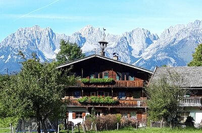 Hof Obernau vor Bergpanorama Traditionelles Holzhaus mit Blumenschmuck vor einer massiven, schneebedeckten Gebirgskette unter blauem Himmel.