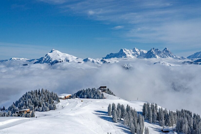 Winterlandschaft am Hahnenkamm Verschneite Berglandschaft mit Skipiste, Gebirgshütten und Nebelmeer vor hohen Alpengipfeln.