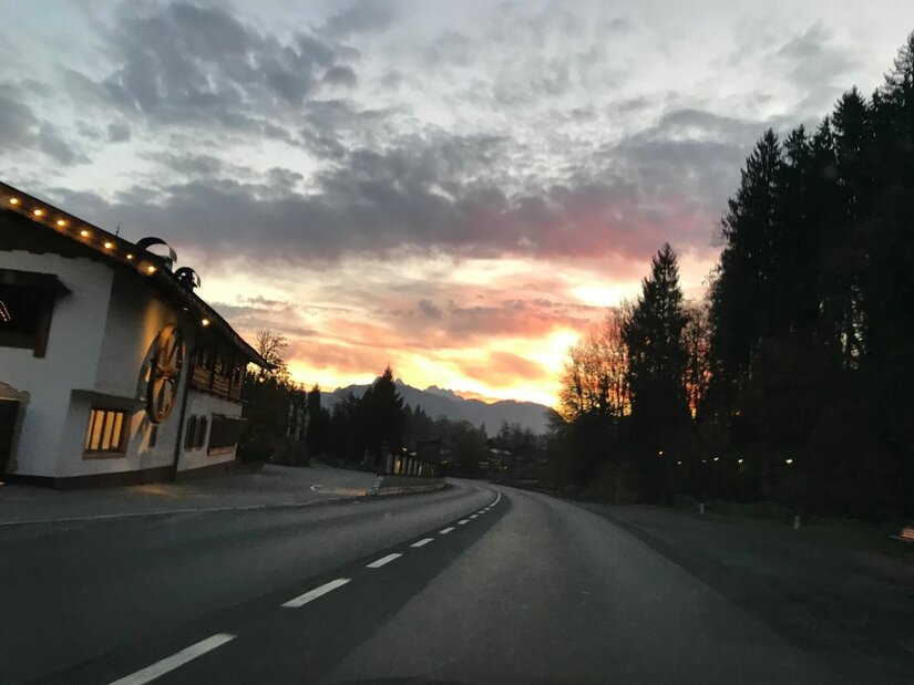 Asphaltierte Landstraße bei Sonnenuntergang mit einem alpinen Gebäude am linken Rand und Wald rechts.