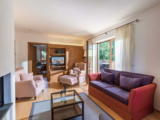 Wohnbereich Appartement Hahnenkamm Wohnzimmer mit rotem Sofa, Sesseln, Holzeinbaumöbeln und Balkonzugang.