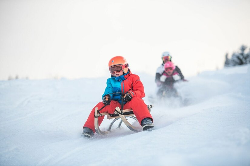 Rodeln am Gaisberg Ein Kind mit Helm und Skibrille fährt auf einem Holzschlitten eine verschneite Piste hinunter.