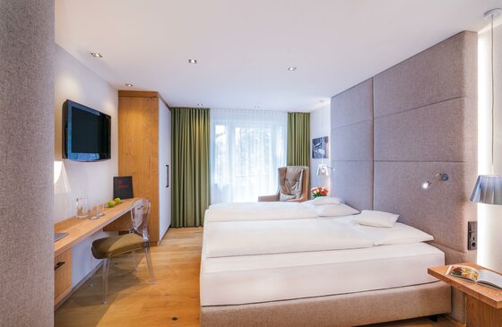Ein modernes Hotelzimmer mit zwei Einzelbetten und freundlicher Einrichtung. Der Raum hat einen Schreibtisch, einen Fernseher und große Fenster, die viel Licht hereinlassen.