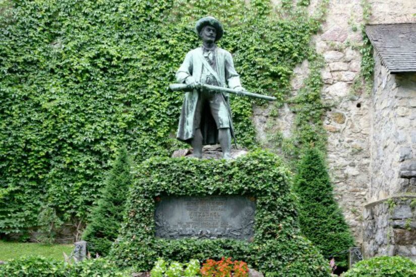 Eine Bronzestatue eines Mannes mit Gewehr steht in einem grünen Garten. Der Hintergrund ist von Efeu und einer Steinmauer umgeben.
