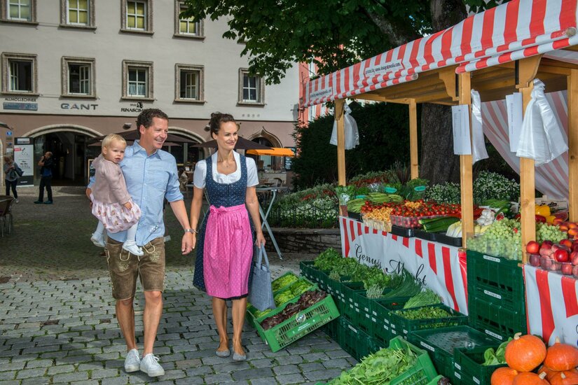 Kitzbüheler Genussmarkt Familie in Tracht besucht einen Marktstand mit frischem Obst und Gemüse auf einem gepflasterten Platz.