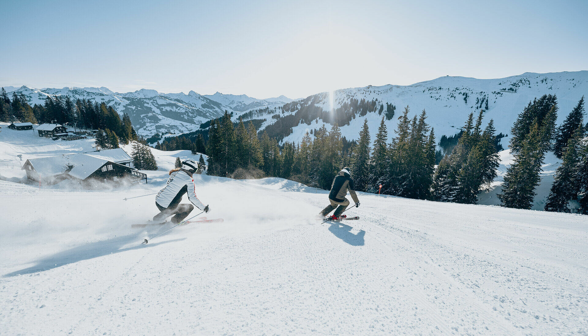 Zwei Skifahrer rasen die Piste hinunter, während die Sonne über den schneebedeckten Gipfeln aufgeht und eine klare Sicht bietet.