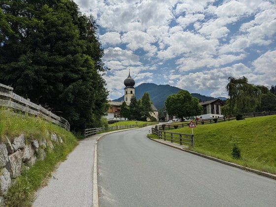 Sonnbergrunde mit Kirchenansicht Asphaltierte Straße führt auf eine Kirche mit Zwiebelturm und bewaldete Berge unter bewölktem Himmel zu.
