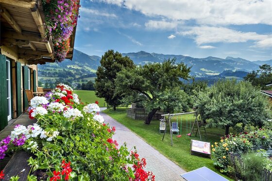 Gartenblick am Lindenhof Blick von einem blumengeschmückten Holzbalkon auf einen Garten mit Spielplatz und Berge im Hintergrund.