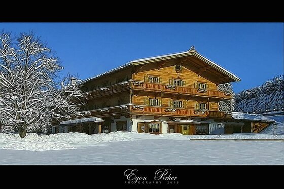Vordergrub im Winter Großes Holzhaus im alpinen Stil mit Balkonen in einer verschneiten Winterlandschaft unter blauem Himmel.