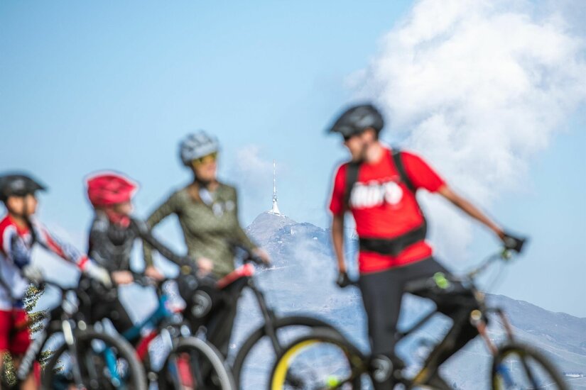 Vier Mountainbiker stehen mit Helmen vor einer Bergkulisse mit Sendeturm.
