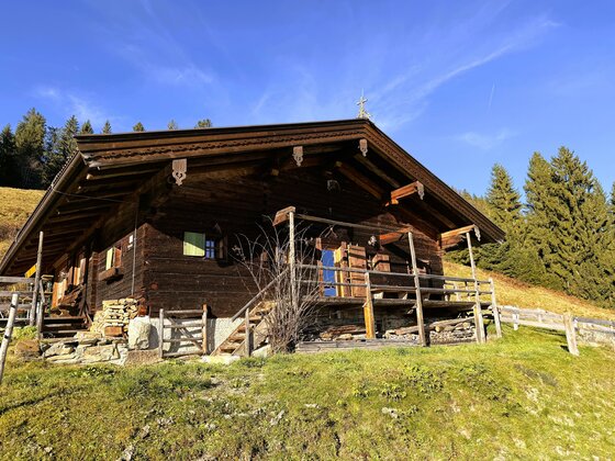 image00005 Eine charmante Almhütte aus Holz, umgeben von üppigem Grün und Bergen. Der Himmel ist blau mit einigen Wolken.